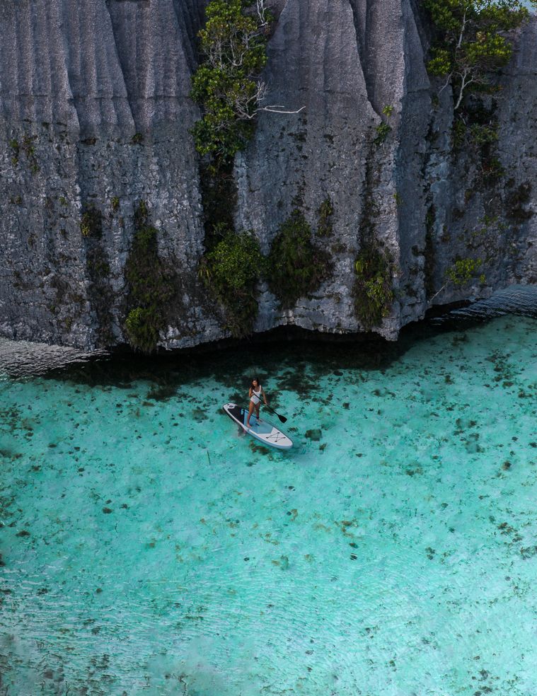Paddleboarder in clear Raja Ampat water beneath limestone cliffs
